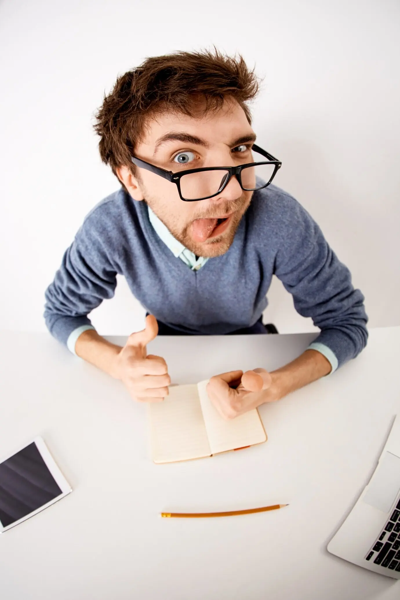 Lustiger, verspielter junger Geschäftsmann mit Brille sitzt am Büroschreibtisch, findet Inspiration, notiert Ideen in einem Notizbuch, streckt die Zunge heraus und zeigt mit dem Finger.