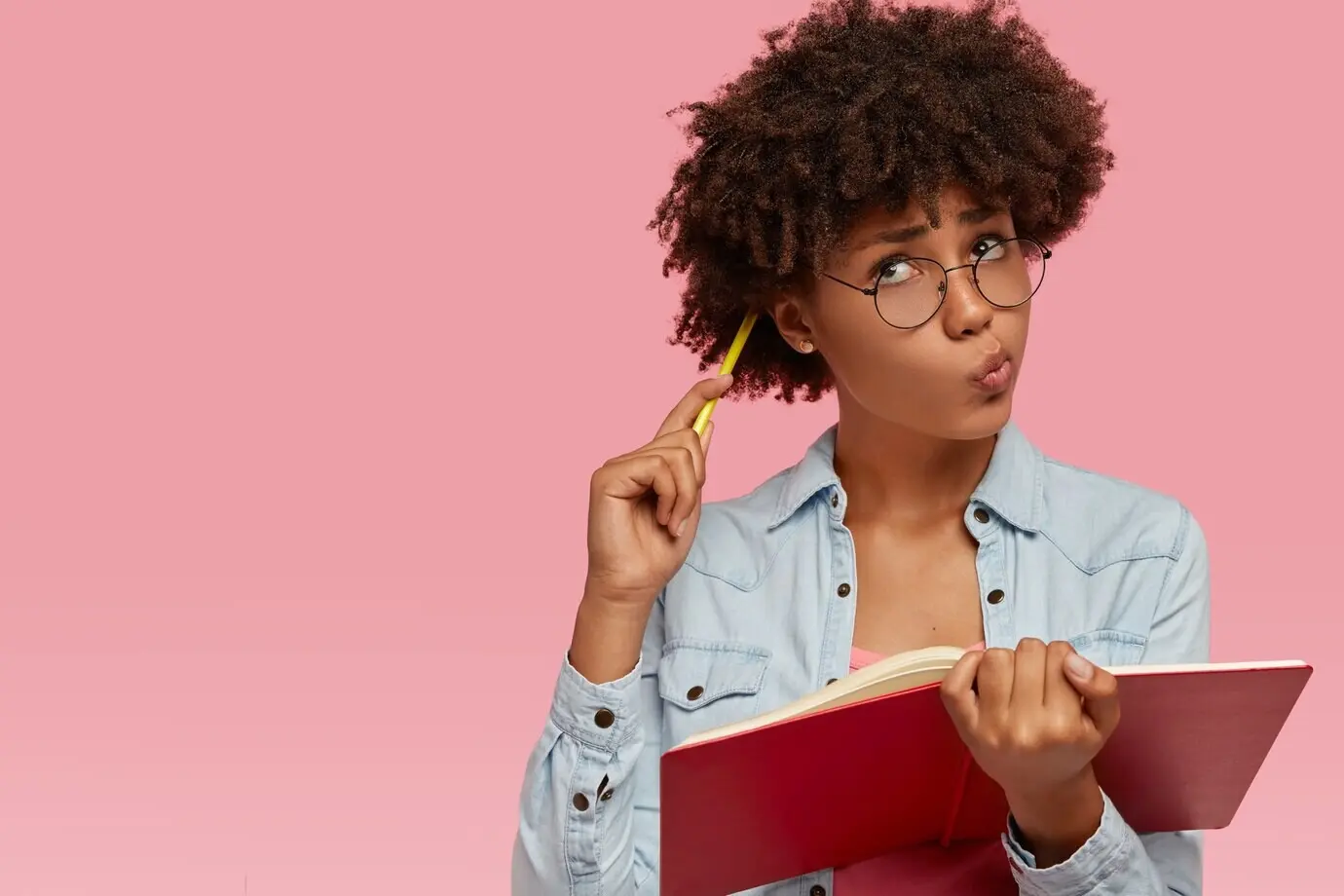 Bild einer nachdenklichen dunkelhäutigen Frau, die sich mit einem Bleistift am Kopf kratzt, die Lippen schürzt und mit zweifelnd-nachdenklichem Ausdruck nach oben blickt, während sie sich kreative Inhalte überlegt und einen Aufsatz für den Unterricht schreibt.