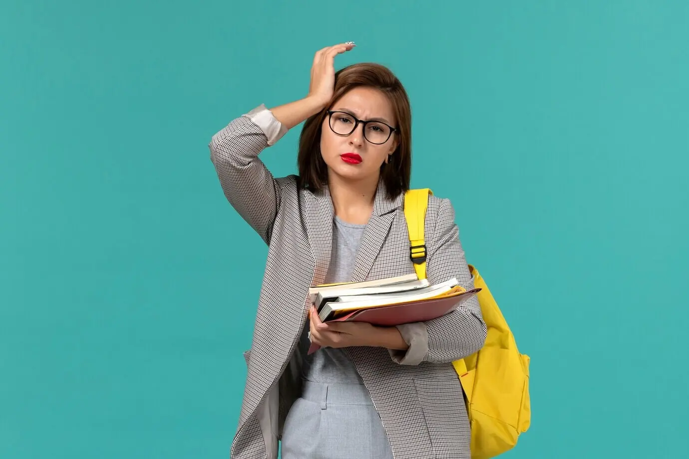 Vorderansicht einer Studentin in grauer Jacke mit gelbem Rucksack, die Bücher hält, vor hellblauer Wand.